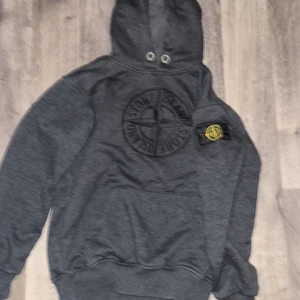 Mörkgrå Stone Island hoodie - Säljer en mörkgrå hoodie från Stone Island med stort broderat logomärke på bröstet och klassisk patch på vänster ärm. Tröjan har huva med metallöljetter, känguruficka och ribbade muddar. Perfekt för dig som gillar streetwear och vill ha något stilrent.