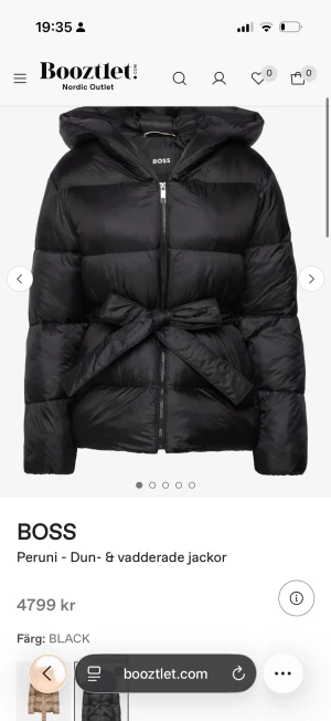 Svart pufferjacka med bälte från BOSS - Snygg svart pufferjacka från BOSS med huva och breda kanaler. Jackan har en cool knytbälte i midjan och dragkedja framtill. Perfekt för kalla dagar och ger en trendig siluett. Materialet är syntet och dun, vilket gör den både varm och lätt.