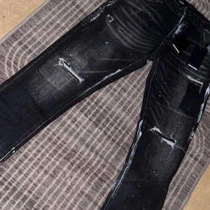 Svarta Dsquared2 jeans med slitningar - Snygga svarta jeans från Dsquared2 med coola slitningar, färgstänk och distressed detaljer. Jeansen har en smal passform och klassisk femficksdesign. Märkeslapp bak och tydliga vita effekter ger en edgy vibe. Perfekta för dig som gillar streetstyle.