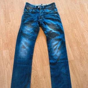 Blå jeans från G-Star Raw Denim - Säljer ett par blå jeans från G-Star Raw Denim i klassisk straight fit. Jeansen har snygga slitningar och kontrastsömmar, fem fickor och knäppning med knappar. Perfekta för dig som gillar en avslappnad men stilren look.