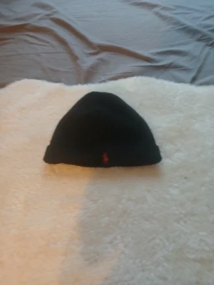 Svart mössa från Polo Ralph Lauren - Svart, klassisk mössa från Polo Ralph Lauren med röd broderad logga framtill. Enkel och stilren design som passar perfekt för kyliga dagar. Mjuk och skön stickad kvalitet som håller dig varm och snygg.