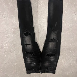 Svarta jeans Supply & Demand - Svarta jeans från Supply & Demand med slitna detaljer och hål på framsidan, samt vita färgstänk för en edgy look. Jeansen har normal passform och klassisk femficksdesign. Perfekta för dig som gillar streetstyle och vill sticka ut.