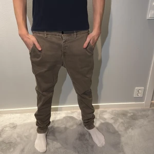 Replay chinos i beige tapered fit - Snygga chinos från Replay i en beige färg med tapered passform. Byxorna har klassiska fickor fram och bak, dragkedja bak och bälteshällor. Materialet är mjukt och skönt bomull, perfekt för en chill och stilren look. Modellen är 186 och väger 70kg
