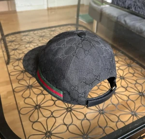 Svart Gucci keps med GG-mönster - Snygg svart keps från Gucci med klassiskt GG-monogram över hela kepsen. Baksidan har justerbart spänne i skinn och en grön-röd rand som ger extra lyxig känsla. Materialet är slitstark bomull och kepsen har en rundad skärm. Perfekt för dig som gillar exklusiva accessoarer.