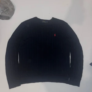 Mörkblå stickad tröja Polo Ralph Lauren - Stilren kabelstickad mörkblå tröja från Polo Ralph Lauren med klassisk rund hals och röd broderad logga på bröstet. Tröjan har lång ärm och ribbade muddar för en snygg passform. Perfekt för lager-på-lager under kyliga dagar.