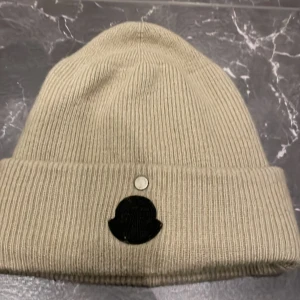 Beige ribbad mössa Moncler x Alyx - Snygg beige ribbstickad mössa från Moncler x 1017 ALYX 9SM. Mössan har en uppvikt kant och svart Moncler-logga framtill. Tillverkad i mjuk ull som håller dig varm och ger en clean look. Perfekt 