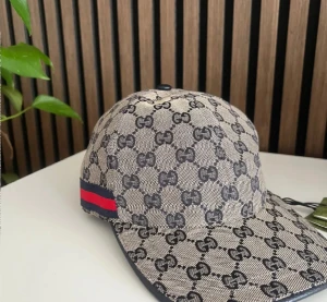 Gucci keps med GG-mönster och röd-blå detalj - Snygg keps från Gucci med klassiskt GG-mönster i mörkgrått på beige bakgrund. Kepsen har en böjd skärm och en röd-blå rand på sidan. Justerbar rem baktill i svart skinn. Perfekt för dig som vill sticka ut med lyxig streetstyle.