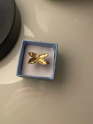 Guldig ring med bladformad detalj - Unik ring i guldton med två stiliserade blad som möts framtill. Ringen har en modern och trendig design som verkligen sticker ut. Perfekt accessoar för dig som vill ha något annorlunda och snyggt på fingret.