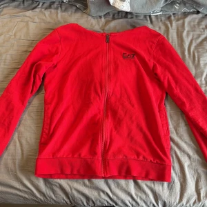 Röd hoodie från EA7 Emporio Armani - Snygg röd hoodie från EA7 Emporio Armani med dragkedja framtill och svart logga på bröstet. Tröjan har huva med snörning och långa ärmar. Tillverkad i mjuk bomull, perfekt för chill eller träning. Cool detalj med svarta ränder på ryggen.