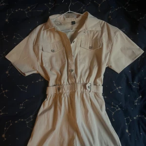 Beige byxdress  - Snygg beige byxdress från SHEIN med krage, korta ärmar och knappar framtill. Den har två bröstfickor med knapp och markerad midja med knappdetaljer. Perfekt för dig som gillar enkel och clean stil.