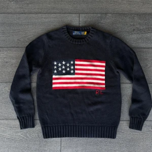 Mörkblå stickad tröja Polo Ralph Lauren - Mörkblå stickad tröja från Polo Ralph Lauren med amerikansk flagga i rött, vitt och blått på bröstet samt RL-broderi. Klassisk rund halsringning och ribbade muddar. Perfekt för dig som gillar preppy och ikonisk stil.
