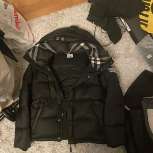 Svart pufferjacka från Burberry - Svart pufferjacka från Burberry med stor huva och klassiskt rutigt foder. Jackan har Burberry London England-tryck på ärmen, dragkedja och knappar framtill samt två fickor med dragkedja. Perfekt för kalla dagar med sin tjocka och quiltade design.