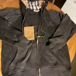 Svart hoodie från Burberry med dragkedja - Svart hoodie från Burberry i storlek M men sitter som s  med klassisk randig insida i huvan. Tröjan har guldfärgade detaljer på snörningen och dragkedja framtill. Mjukt material och ribbade muddar vid ärmslut och nederkant. Perfekt för dig som gillar stilrena märkesplagg.
