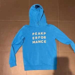 Blå hoodie från Peak Performance - Snygg blå hoodie från Peak Performance med vit text på bröstet. Tröjan har huva, lång ärm och en stor magficka framtill. Tillverkad i mjuk bomull som är skön mot huden. Perfekt för dig som gillar sportig och avslappnad stil.