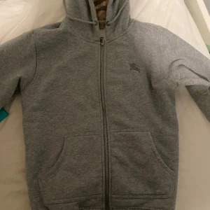 Grå hoodie från Burberry med dragkedja - Snygg grå hoodie från Burberry i storlek M med klassisk dragkedja framtill och huva med ikoniskt rutigt foder. Tröjan har två stora fickor och ett diskret Burberry-logotryck på bröstet. Perfekt för dig som gillar stilrena och exklusiva plagg.