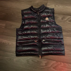 Mörkblå dunväst från Moncler - Snygg mörkblå dunväst från Moncler med glansig finish och klassisk logga på bröstet. Västen har hög krage, två dragkedjefickor och stängs med dragkedja framtill. Ända negativa är att man behöver ta på sig den som en hoodie för dragkedjan krånglar nere