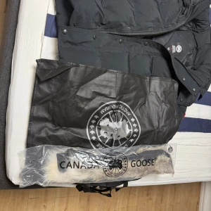 Svart dunjacka från Canada Goose M - Säljer en svart dunjacka från Canada Goose i storlek M. Jackan har huva, dragkedja och knappar framtill, samt klassisk patch på ärmen. Medföljer avtagbar pälskant till huvan. Perfekt för kalla vinterdagar och riktigt snygg streetstil.