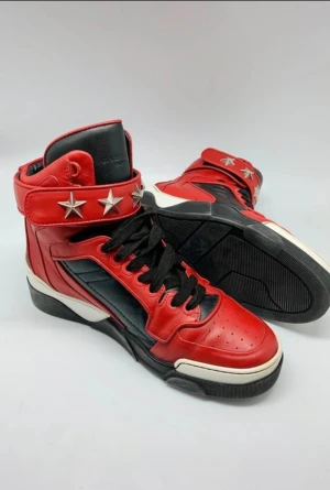 Givenchy röda high-top sneakers - Säljer ett par grymma high-top sneakers från Givenchy i rött och svart skinn med coola silverfärgade stjärndetaljer runt ankeln. Skorna har svarta snören, perforerad tå och vit sula med svart kontrast. Perfekt för dig som vill sticka ut med lyxig streetstyle.