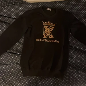 Svart Dolce & Gabbana sweatshirt M - Svart sweatshirt från Dolce & Gabbana i storlek M med guldigt tryck på bröstet och en stor K med krona. Tröjan har rund hals, långa ärmar och ribbade muddar. Materialet är mjukt och känns som bomull, perfekt för dig som gillar exklusiva märken och snygg design.