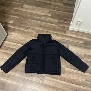 Svart pufferjacka från Bik Bok XS - Svart pufferjacka från Bik Bok i storlek XS. Jackan har hög krage, raka långa ärmar och två fickor med knappstängning. Materialet är återvunnet polyester och jackan stängs med dragkedja framtill. Perfekt för kalla dagar med sin quiltade design.