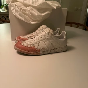 Maison Margiela vita sneakers med färgstänk - Maison Margiela sneakers i vitt med unika röda färgstänk på tå och sidor. Klassisk låg modell med snörning och rund tå. Snyggt textilmaterial med skinnpaneler och platt sula. Perfekt för dig som gillar statement pieces och exklusiv design. Storlek 40 men passar 41. Innersula är 26cm