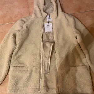 Beige/gräddvit mamma/gravid/amning/teddyjacka med huva XL - Oanvänd/ny m lapp kvar (nypris 649:-) Mysig teddyjacka från Mamalicious i beige med huva och dragkedja framtill. Jackan har två fickor och är tillverkad i fluffigt fleece-material som ger en chill vibe. Fodrad o varm. Perfekt för dig som vill ha det varmt och skönt. Lätt tvätta! Jackan går även att ha efter graviditet o då bara att ta bort den extra infällda delen fram o använda m blixtlåset som vanligt. P g a aprikos bakgrund ser jackan mer aprikos ut men den är gräddvit/äggskal/beige