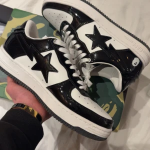 bape skor - Säljer ett par BAPE sneakers i svart och vitt med den ikoniska stjärnan på sidan. Skorna har snörning, platt sula och rund tå. Materialet är blankt skinn som ger en riktigt clean och modern vibe. Perfekt för dig som vill sticka ut med streetwear.