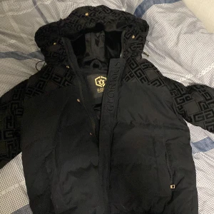 Svart pufferjacka Glorious Gangsta - Svart pufferjacka från Glorious Gangsta med fet logga på insidan och mönstrade partier på huva och ärmar. Jackan har dragkedja, guldiga detaljer och en stor huva. Ny kostar runt 1500, Köpt förra vintern inga fel på varan 😃