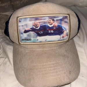 Beige och marinblå keps från Diverzo - Limited Edition keps från Diverzo med beige manchester framtill och marinblå mesh baktill. Framsidan har en stor patch med fotbollsmotiv och broderad ram. Justerbar rem bak i svart skinn. Perfekt för dig som gillar sportig stil och unika detaljer.