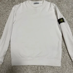 Vit sweatshirt från Stone Island - Vit långärmad sweatshirt från Stone Island med klassisk rund hals och ikonisk logotyp-patch på vänster ärm. Tröjan har en clean look och är tillverkad i mjuk bomull som är skön att bära. Perfekt för dig som gillar stilrena och trendiga plagg.