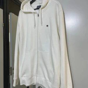 Vit hoodie från Champion med dragkedja - Vit hoodie från Champion med klassisk logga på bröstet. Tröjan har huva med snörning, dragkedja framtill och två stora fickor. Mjuk bomullskänsla och långärmad modell, perfekt för chill dagar eller när du vill ha en clean look.