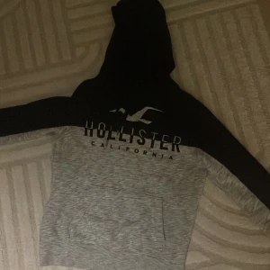 Svart och grå hoodie från Hollister - Snygg hoodie från Hollister i storlek S med svart överdel och huva samt gråmelerad nedre del. Stor Hollister-logga och texten 'California' på bröstet. Tröjan har en stor magficka och långa ärmar, perfekt för chill dagar.