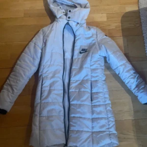Ljusgrå lång dunjacka från Nike - Säljer en lång, ljusgrå dunjacka från Nike med huva och svart logga på bröstet. Jackan är quiltad och har dragkedja framtill samt tryckknappar vid halsen. Perfekt för kalla dagar och har en clean, sportig look.