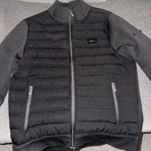 Svart pufferjacka från Stone Island - Svart pufferjacka från Stone Island med quiltad framsida och två dragkedjeförsedda fickor. Jackan har ribbad krage och muddar, samt en snygg logotyp på bröstet. Materialet är polyester och jackan har en normal passform med långärmade ärmar.