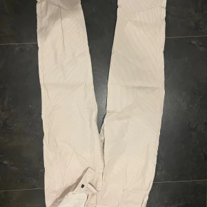 Randiga kostymbyxor från Mango - Snygga kostymbyxor från Mango i ljusbeige med tunna vertikala ränder. Byxorna har klassisk passform, bälteshällor och stängs med knapp och dragkedja. Perfekta för en clean och stilren look. Tillverkade i bomull för en skön känsla.