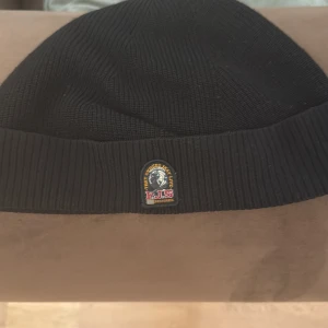 Svart ribbad mössa från Parajumpers - Svart ribbstickad mössa från Parajumpers med klassisk logopatch framtill. Snygg och enkel design som funkar till alla jackor. Mjuk och varm stickad kvalitet, perfekt för kyliga dagar. Äkta köpte från NK finns Scan 