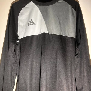 Svart och grå långärmad Adidas målvaktströja - Säljer en svart och grå långärmad målvaktströja från Adidas med Climalite-material. Tröjan har vadderade armbågar för extra skydd och ett stilrent blockmönster upptill. Adidas-loggan är tryckt på bröstet. Perfekt för dig som vill ha en snygg och funktionell tröja på planen.
