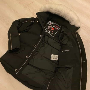 Svart dunjacka Moose Knuckles med päls - Svart dunjacka från Moose Knuckles med fluffig vit päls på huvan, silverfärgade dragkedjor och knappar. Jackan har lång ärm, flera fickor och coola detaljer på axlarna. Perfekt för kalla dagar och har en snygg, urban look.