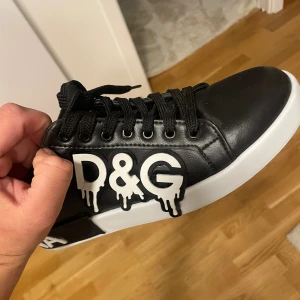 Svarta sneakers från Dolce & Gabbana - Säljer ett par svarta sneakers från Dolce & Gabbana med vit sula och coolt D&G-tryck i drip-style på sidan. Skorna har snörning, vita detaljer vid hälen och är tillverkade i skinn. Perfekta för dig som vill sticka ut med en unik streetstyle.