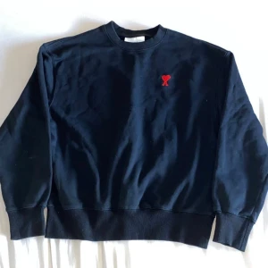 Svart sweatshirt från Ami med rött hjärta - Svart sweatshirt från Ami Alexandre Mattiussi med klassisk rund hals och lång ärm. På bröstet sitter ett broderat rött hjärta med bokstaven A. Tröjan är gjord i mjuk bomull och har ribbade muddar vid ärmslut och nederkant. Perfekt för en chill och stilren look.