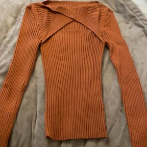 Rostbrun ribbad omlott-tröja - Säljer en snygg rostbrun långärmad tröja med ribbad struktur och omlott-detalj vid halsen. Tröjan har en tight passform och är perfekt för dig som gillar trendiga och unika plagg. Materialet känns mjukt och stretchigt, vilket gör den skön att bära. Pris kan diskuteras och köparen står för frakt