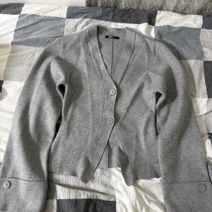 Slutsåld Grå kofta från Gina Tricot - Snygg grå kofta från Gina Tricot med v-ringad hals och stora knappar framtill. Modellen är croppad och har långa ärmar med knappdetalj vid mudden. Mjuk stickad kvalitet som är perfekt för lager-på-lager under hösten och vintern.