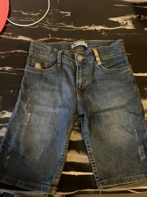 Blå jeansshorts från Burberry - Säljer ett par klassiska blå jeansshorts från Burberry med snygga detaljer som Burberrys rutiga tyg på fickan och bältet. Shortsen har normal passform, fem fickor och knappstängning framtill. Perfekt för dig som gillar stilrena och tidlösa plagg med designer-touch.