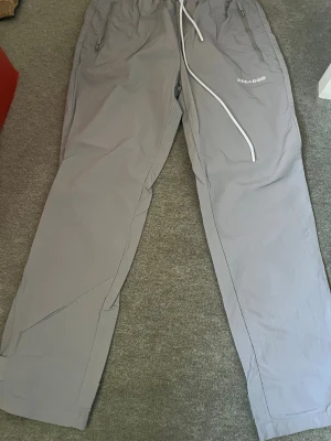 Grå Pegador byxor med snörning XS - Säljer ett par grå byxor från Pegador i storlek XS. De har elastisk midja med vit snörning, dragkedjeförsedda fickor och rakt snitt. Materialet är lätt och syntetiskt, perfekt för en chill och avslappnad stil.