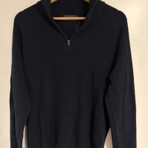 Mörkblå stickad tröja Dressmann - Tröjan är helt oanvänd. Snygg mörkblå stickad tröja från Dressmann Premium med half zip och hög krage. Tröjan är ribbad och har långärmad passform, perfekt för dig som gillar stilrena och klassiska plagg. Materialet är mjukt och håller värmen under kyliga dagar.