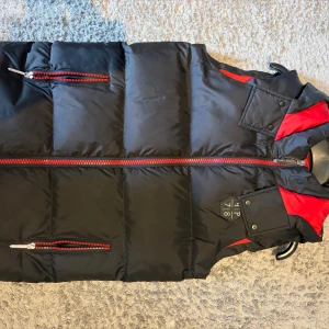 philipp plein väst - Snygg svart dunväst från 4IP 718 med röda detaljer och dragkedjor. Västen har en huva, två fickor med röda dragkedjor och ett coolt emblem på bröstet. Perfekt för dig som vill ha en sportig och modern look när det är kyligt ute.