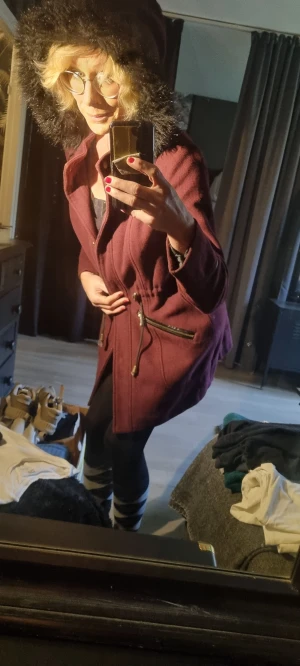 Vinröd kappa med fuskpäls från Atmosphere - Säljer en vinröd kappa från Atmosphere med svart fuskpäls på huvan. Jackan har dragkedjor vid fickorna, snörning i midjan och snygga detaljer i guld. Perfekt för kalla dagar och ger en riktigt stilren look.