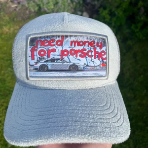 Grå keps från Revenite med Porsche-tryck - Snygg grå keps från Revenite med mesh på baksidan och justerbar snapback. Framtill finns ett stort patch med texten 'need money for Porsche' och en bild på en Porsche framför graffitivägg. Perfekt för dig som gillar streetstyle och bilar. (Limited edition) (Finns bara 500 av den) original kartong finns inte. helt ny aldrig använd