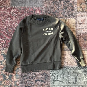 Mörkgrön sweatshirt från GANT - Snygg mörkgrön sweatshirt från GANT med tryck på bröstet där det står 'GANT USA EST. 1949 NEW HAVEN'. Tröjan har rund hals, ribbade muddar och är långärmad. Perfekt för dig som gillar klassisk och sportig stil.
