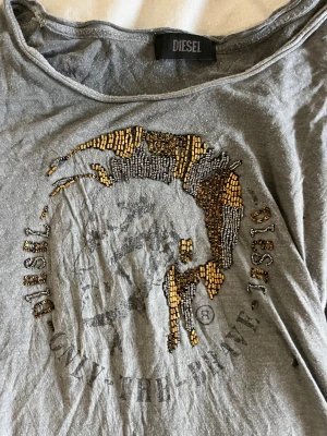 Grå långärmad tröja från Diesel - Snygg grå långärmad tröja från Diesel med bred halsringning och coolt paljettbroderat motiv framtill i guld och silver. Tröjan är i mjuk bomull och har ett avslappnat, lite loose fit. Perfekt för dig som gillar statement-plagg och streetstyle.